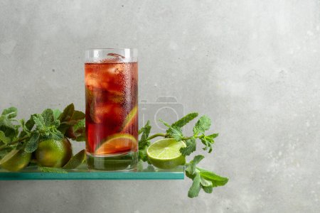 Téléchargez les photos : Cocktail Cuba Libre avec glace, rhum brun, cola, menthe et citron vert. Boisson glacée dans un verre embrumé sur fond de béton gris. Espace de copie. - en image libre de droit