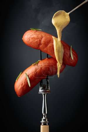 Foto de Boiled sausages with rosemary and mustard on a fork. Hot sausages with smoke on a black background. - Imagen libre de derechos