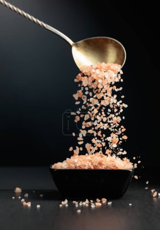 Téléchargez les photos : Le sel rose est versé dans un petit bol noir. Sel himalayen sur fond noir. - en image libre de droit