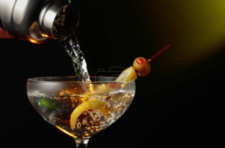 Foto de Martini con aceitunas verdes y limón. El cóctel se vierte de una coctelera en un vaso. - Imagen libre de derechos