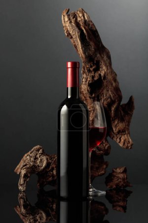 Photo for Glass and bottle of red wine. In the background old weathered snag. Black reflective background. Frontal view with space for your text. - Royalty Free Image