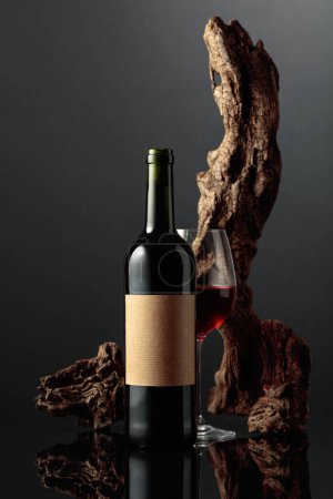Photo for Glass and bottle of red wine with old empty label. In the background old weathered snag. Black reflective background. Frontal view with space for your text. - Royalty Free Image