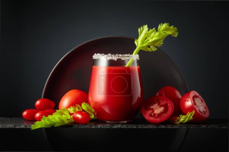 Foto de Jugo de tomate fresco con apio y un montón de verduras maduras sobre un fondo negro. Vidrio adornado con sal marina. - Imagen libre de derechos