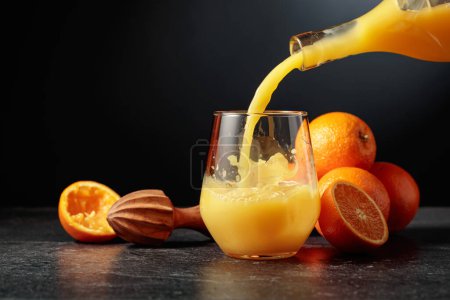 Foto de Verter el jugo de naranja de una botella en un vaso con hielo. Fondo negro con espacio de copia. - Imagen libre de derechos