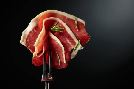 Photo for Sliced prosciutto with rosemary on a fork, black background, copy space. - Royalty Free Image