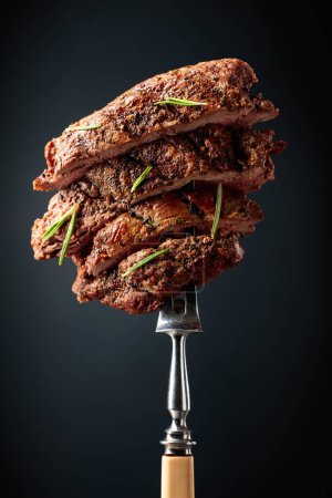 Téléchargez les photos : Bifteck de bœuf côtelé grillé avec romarin sur fond noir. Steak de boeuf sur une fourchette saupoudrée de romarin. - en image libre de droit