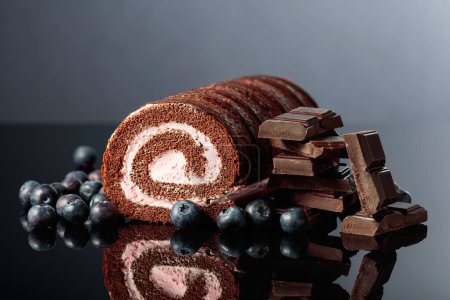 Foto de Chocolate roll cake with blueberries and a broken black chocolate bar on a black reflective background. - Imagen libre de derechos