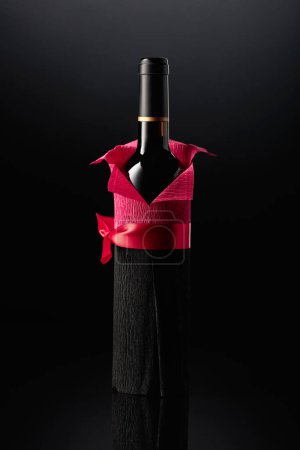 Téléchargez les photos : Bottle of red wine wrapped in crepe paper on a black background. The bottle looks like a man in a red shirt. - en image libre de droit