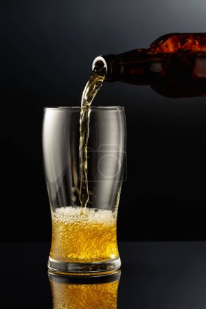 Foto de Verter cerveza de una botella en un vaso. Vaso de cerveza sobre fondo negro reflectante. - Imagen libre de derechos