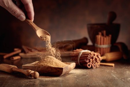 Photo for Cinnamon powder is poured into a wooden bowl. In the background are kitchen utensils and cinnamon sticks. - Royalty Free Image