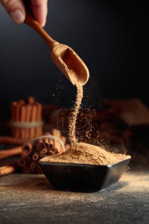 Photo for Cinnamon powder is poured into a black bowl. In the background are kitchen utensils and cinnamon sticks. - Royalty Free Image