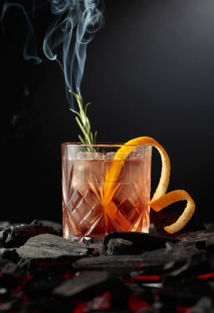 Foto de Cóctel a la antigua con hielo, cáscara de naranja y romero. Whisky con ramita ardiente de romero sobre fondo oscuro. - Imagen libre de derechos