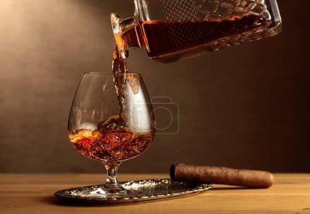 Foto de Brandy is poured from a decanter into a snifter glass. Cognac and cigar on an oak table. - Imagen libre de derechos