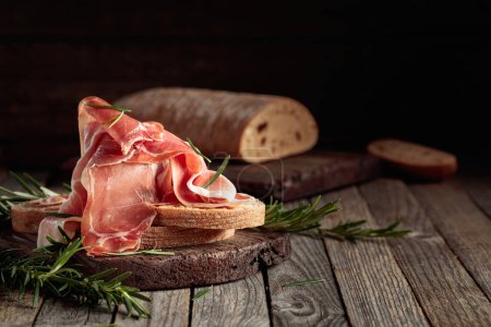 Photo for Prosciutto with bread and rosemary on an old wooden table. Copy space. - Royalty Free Image