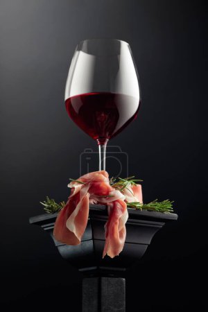 Photo for Glass of red wine and prosciutto with rosemary on a black podium. - Royalty Free Image