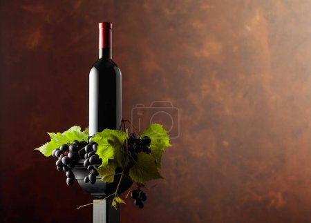 Foto de Botella de rojo en un podio negro. Vino con uvas azules y ramas de vid. Fondo marrón con espacio de copia. - Imagen libre de derechos