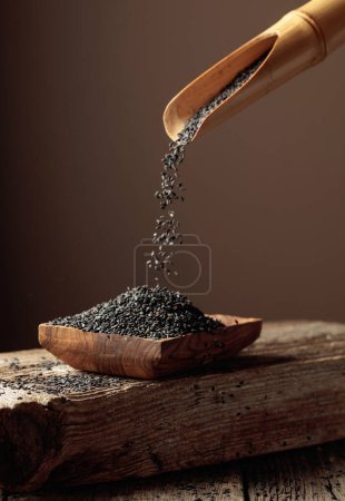 Téléchargez les photos : Les grains de sésame noir sont versés avec une cuillère en bois dans le plat en bois. Espace de copie. - en image libre de droit