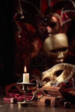 Téléchargez les photos : Bougie allumée, dés et masques de carnaval sur une vieille table en bois. - en image libre de droit