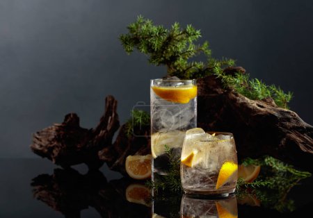 Foto de Cóctel gin-tonic con hielo y limón sobre fondo negro reflectante. Bebida helada con viejos snags y ramas de enebro. - Imagen libre de derechos