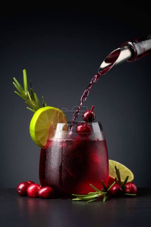 Photo for Cranberry cocktail garnished with berries, lime, and rosemary. In frozen glass with ice is pouring cranberry liquor. - Royalty Free Image