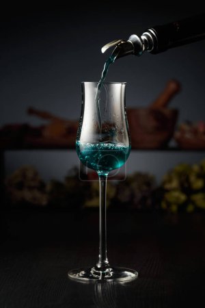 Téléchargez les photos : Le gin bleu est versé d'une bouteille vintage dans un verre. En arrière-plan herbes séchées et vieux ustensiles de cuisine. - en image libre de droit
