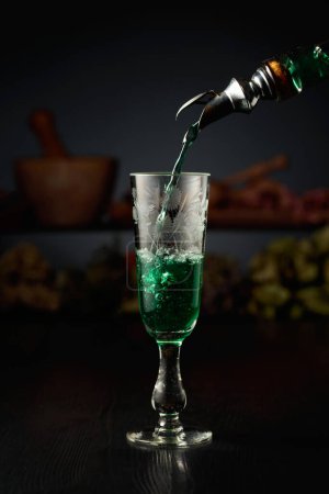 Téléchargez les photos : La liqueur verte aux herbes est versée d'une bouteille vintage dans un verre. En arrière-plan herbes séchées et vieux ustensiles de cuisine. - en image libre de droit