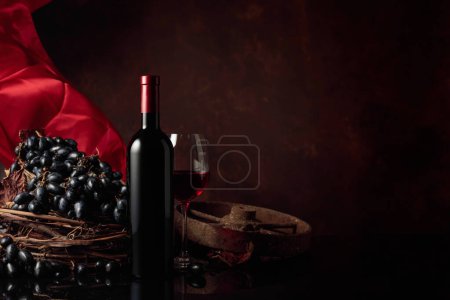 Téléchargez les photos : Verre et bouteille de vin rouge avec raisins sur fond noir réfléchissant. Rideau en satin rouge flotte dans le vent. Espace de copie. - en image libre de droit