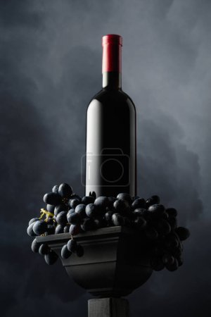 Photo for Blue grape and a bottle of red wine on a background of cloudy sky. Side and bottom view. Selective focus. - Royalty Free Image