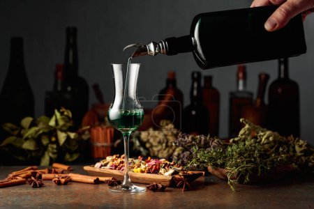 Téléchargez les photos : La liqueur verte aux herbes est versée d'une bouteille vintage dans un verre. Sur une table des herbes séchées, des fleurs, des épices et de vieux ustensiles de cuisine. Concept de phytothérapie. - en image libre de droit