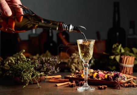 Téléchargez les photos : La liqueur ou le mélange à base de plantes jaunes est versé d'une bouteille vintage dans un verre. Sur une table des herbes séchées, des fleurs, des épices et de vieux ustensiles de cuisine. Concept de phytothérapie. - en image libre de droit