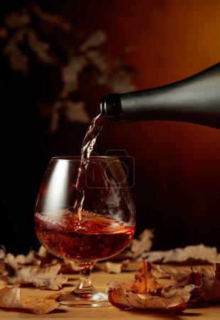Téléchargez les photos : Le brandy est versé d'une bouteille dans un verre. Un soupçon de brandy sur une vieille table de chêne avec des feuilles de chêne séchées. - en image libre de droit