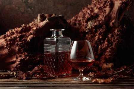 Foto de Decantador y copa de brandy sobre una vieja mesa de madera. En el fondo hay viejos enganches y hojas de vid secas. Copiar espacio. - Imagen libre de derechos
