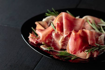 Photo for Italian prosciutto or Spanish jamon with rosemary on a black plate. - Royalty Free Image