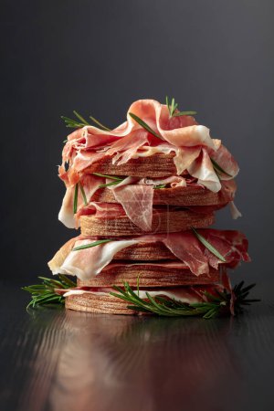 Photo for Italian prosciutto or Spanish jamon with bread and rosemary on a black wooden table. Copy space. - Royalty Free Image