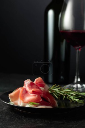 Foto de Prosciutto con romero y vino tinto sobre fondo negro. Copia espacio para tu texto. - Imagen libre de derechos