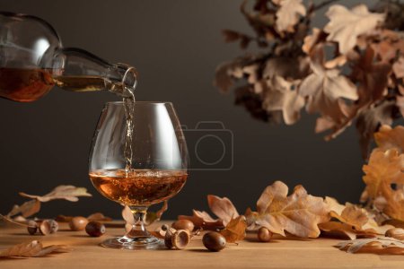 Photo for Brandy is poured from a bottle into a glass. Snifter of cognac on a wooden table with dried-up oak leaves. - Royalty Free Image