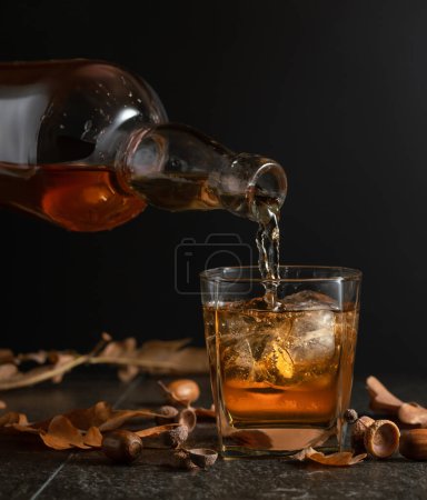 Foto de El whisky se vierte de una botella en un vaso con hielo. Whisky en vaso congelado con hielo sobre una mesa negra con hojas secas de roble. - Imagen libre de derechos