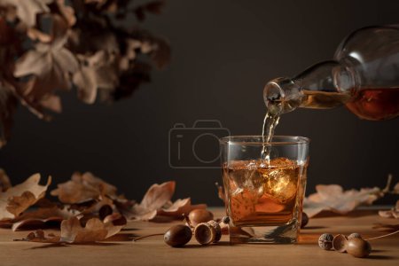 Foto de El whisky se vierte de una botella en un vaso con hielo. Whisky en vidrio congelado con hielo sobre una mesa de madera con hojas secas de roble. - Imagen libre de derechos