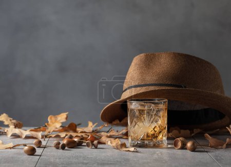 Téléchargez les photos : Whisky avec glace sur une table en pierre avec des feuilles de chêne séchées et un chapeau d'homme. - en image libre de droit