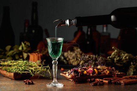 Téléchargez les photos : La liqueur verte aux herbes est versée d'une bouteille vintage dans un verre. Sur une table des herbes séchées, des fleurs, des épices et de vieux ustensiles de cuisine. Concept de phytothérapie. - en image libre de droit