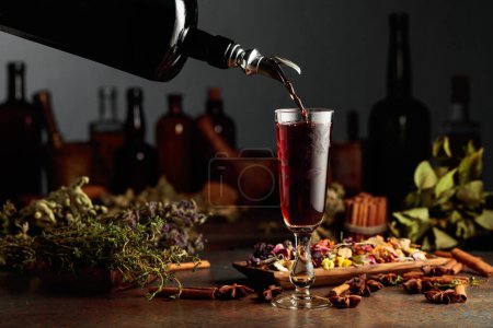 Téléchargez les photos : La liqueur ou le baume brun à base de plantes est versé d'une bouteille vintage dans un verre. Sur une table des herbes séchées, des fleurs, des épices et de vieux ustensiles de cuisine. Concept de phytothérapie. - en image libre de droit