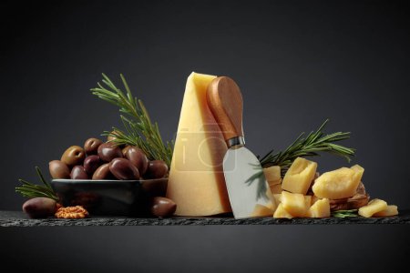 Foto de Queso parmesano con cuchillo, aceitunas y romero sobre fondo negro. - Imagen libre de derechos