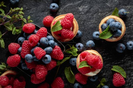 Photo for Small tartlets with fresh raspberries and blueberries garnished with mint on a black background. Top view. - Royalty Free Image