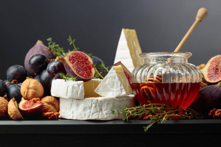 Foto de Queso Camembert con higos, uvas, nueces, miel y tomillo. Queso suave con frutas sobre fondo negro. - Imagen libre de derechos