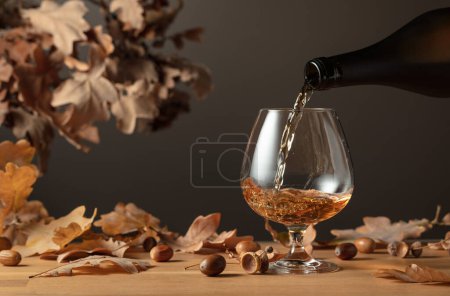 Foto de El brandy se vierte de una botella en un vaso. Francotirador de coñac sobre una mesa de madera con hojas secas de roble. - Imagen libre de derechos