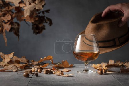 Foto de Francotirador de brandy en una mesa de piedra con hojas secas de roble y sombrero de hombre. - Imagen libre de derechos