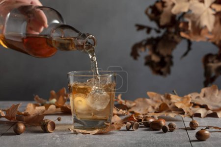 Photo for Whiskey is poured from a bottle into a glass with ice. Whiskey in frozen glass with ice on a grey stone table with dried-up oak leaves. - Royalty Free Image
