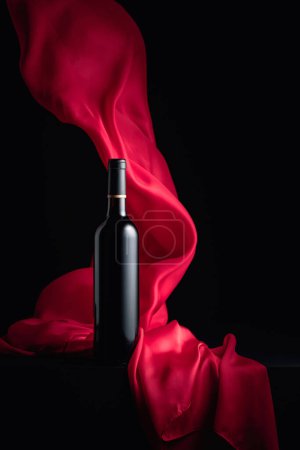 Téléchargez les photos : Bouteille de vin rouge et volutes de tissu rouge sur fond noir. - en image libre de droit