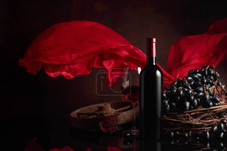 Téléchargez les photos : Verre et bouteille de vin rouge avec raisins sur fond noir réfléchissant. Rideau en satin rouge flotte dans le vent. Espace de copie. - en image libre de droit