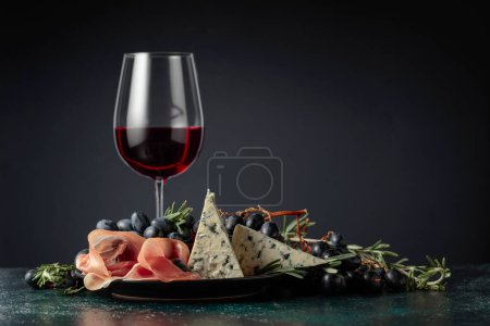 Photo for Red wine with grapes, rosemary, prosciutto, and blue cheese on a dark background. - Royalty Free Image